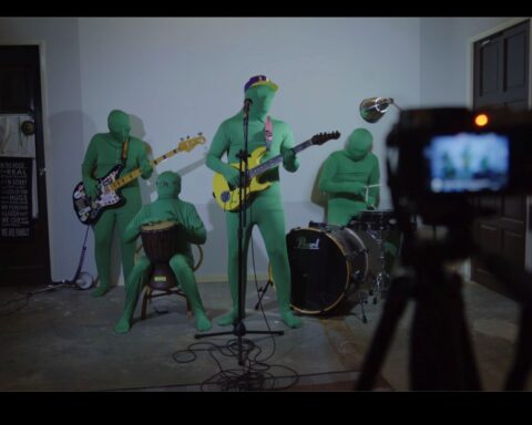 A punk band of four members performs on stage while a camera is in the foreground filming them. The musicians are dressed completely in green suits with their bodies and faces fully covered.