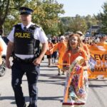 A Mohawk woman in an orange shirt bangs a drum and leads a march. She is walking beside a uniformed police officer.