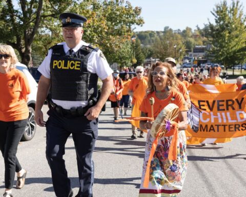 A Mohawk woman in an orange shirt bangs a drum and leads a march. She is walking beside a uniformed police officer.
