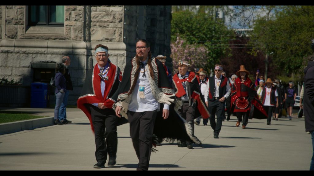 A series of men in Haida regalia walk outside a courthouse.