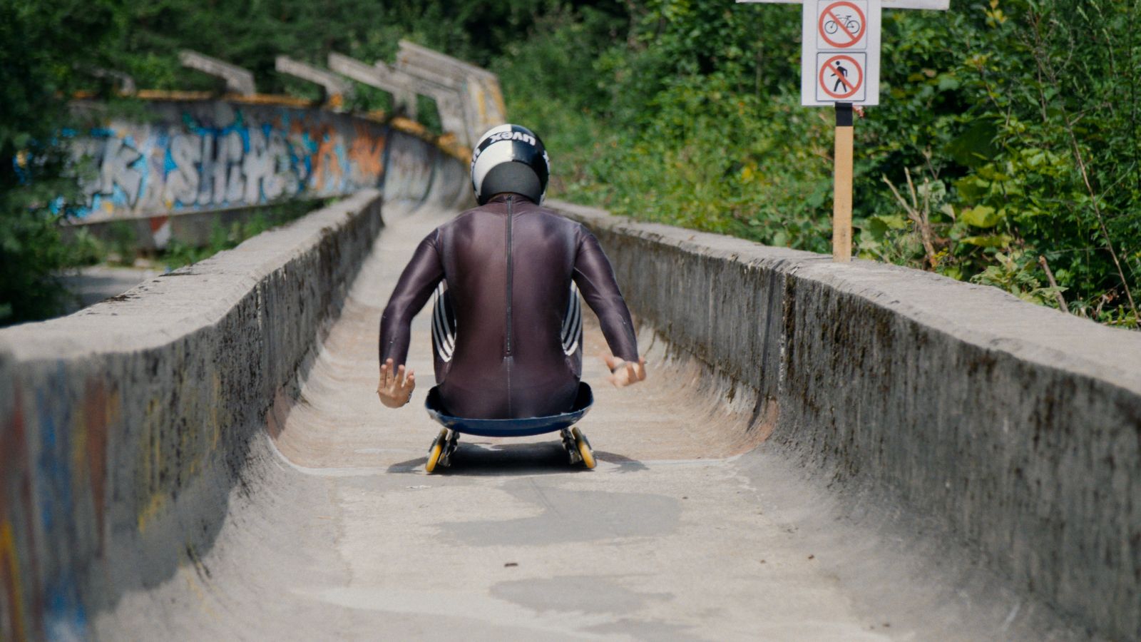 View from behind as a luge player slides down a practice track. There is no snow and he is gliding down bare concrete. He is wearing a black uniform and a white helmet.