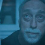 An older Caucasian man with a white beard and moustache looks up to the sky through his window. The image has a blue tint.