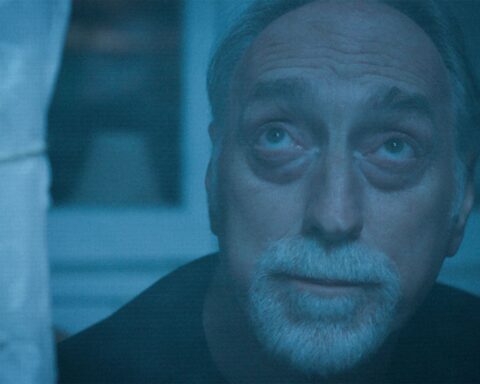 An older Caucasian man with a white beard and moustache looks up to the sky through his window. The image has a blue tint.