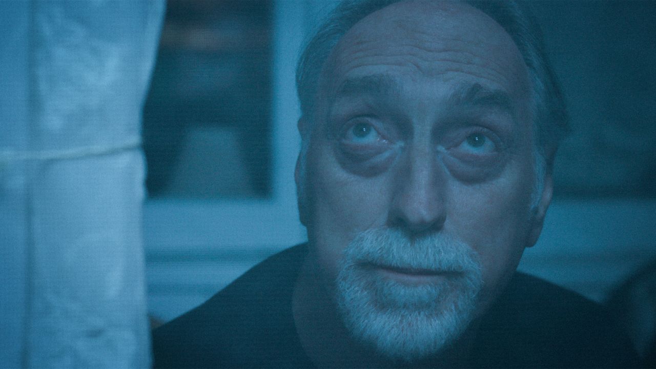 An older Caucasian man with a white beard and moustache looks up to the sky through his window. The image has a blue tint.