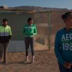 Three Venezuelans stand on a sandy patch of ground before a fence. They are all looking screen right. It is a sunny day.