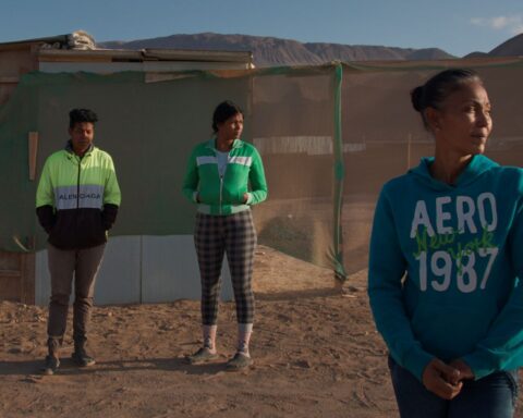 Three Venezuelans stand on a sandy patch of ground before a fence. They are all looking screen right. It is a sunny day.