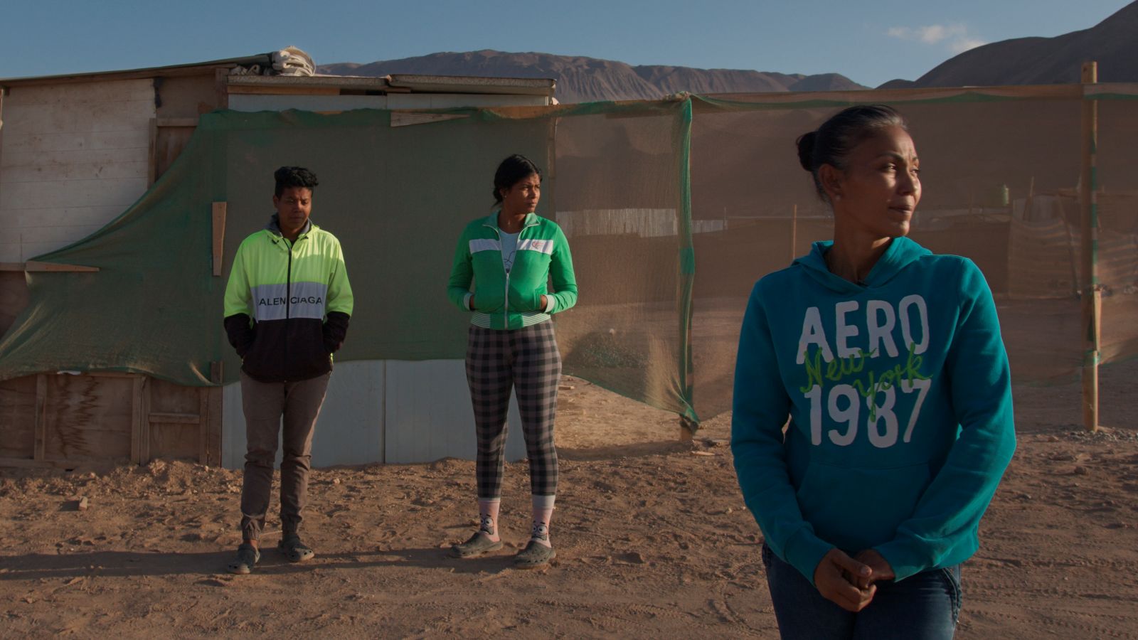 Three Venezuelans stand on a sandy patch of ground before a fence. They are all looking screen right. It is a sunny day.