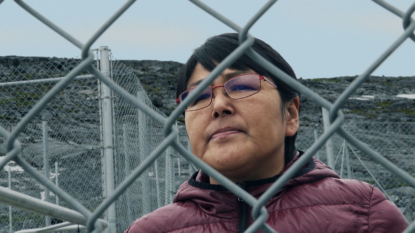 A close-up shot of a woman standing on the other side of a wire fence. She is wearing a burgundy winter jacket and eyeglasses, looking at what is on the other side of the fence.