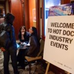 A white sign with black text reads Welcome to Hot Docs Industry Events. There a blue and purple top border on the sign. Three people are talking in the background, appearing in a busy hotel.