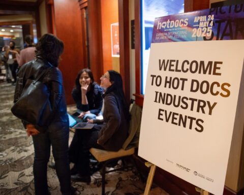 A white sign with black text reads Welcome to Hot Docs Industry Events. There a blue and purple top border on the sign. Three people are talking in the background, appearing in a busy hotel.
