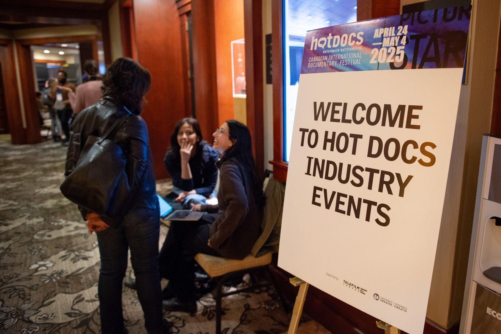 A white sign with black text reads Welcome to Hot Docs Industry Events. There a blue and purple top border on the sign. Three people are talking in the background, appearing in a busy hotel.