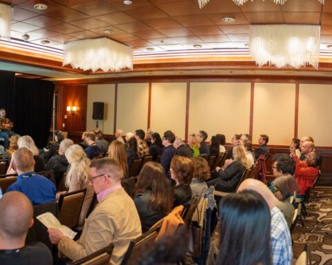 A crowded audience sits in a conference room where four panelists discuss a topic. The screen behind them advertises the topic of "Truth or Bias".