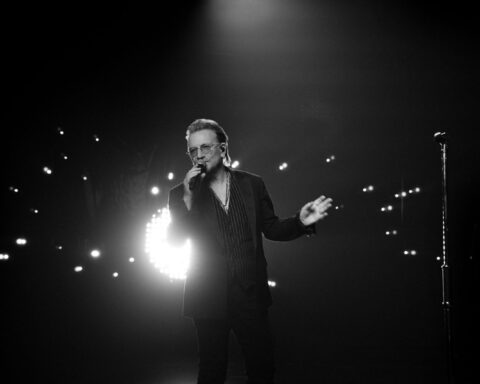 A black and white image of singer Bono standing on stage. He is wearing a black suite with sunglasses, and holding a microphone. There is a series of lights behind him.