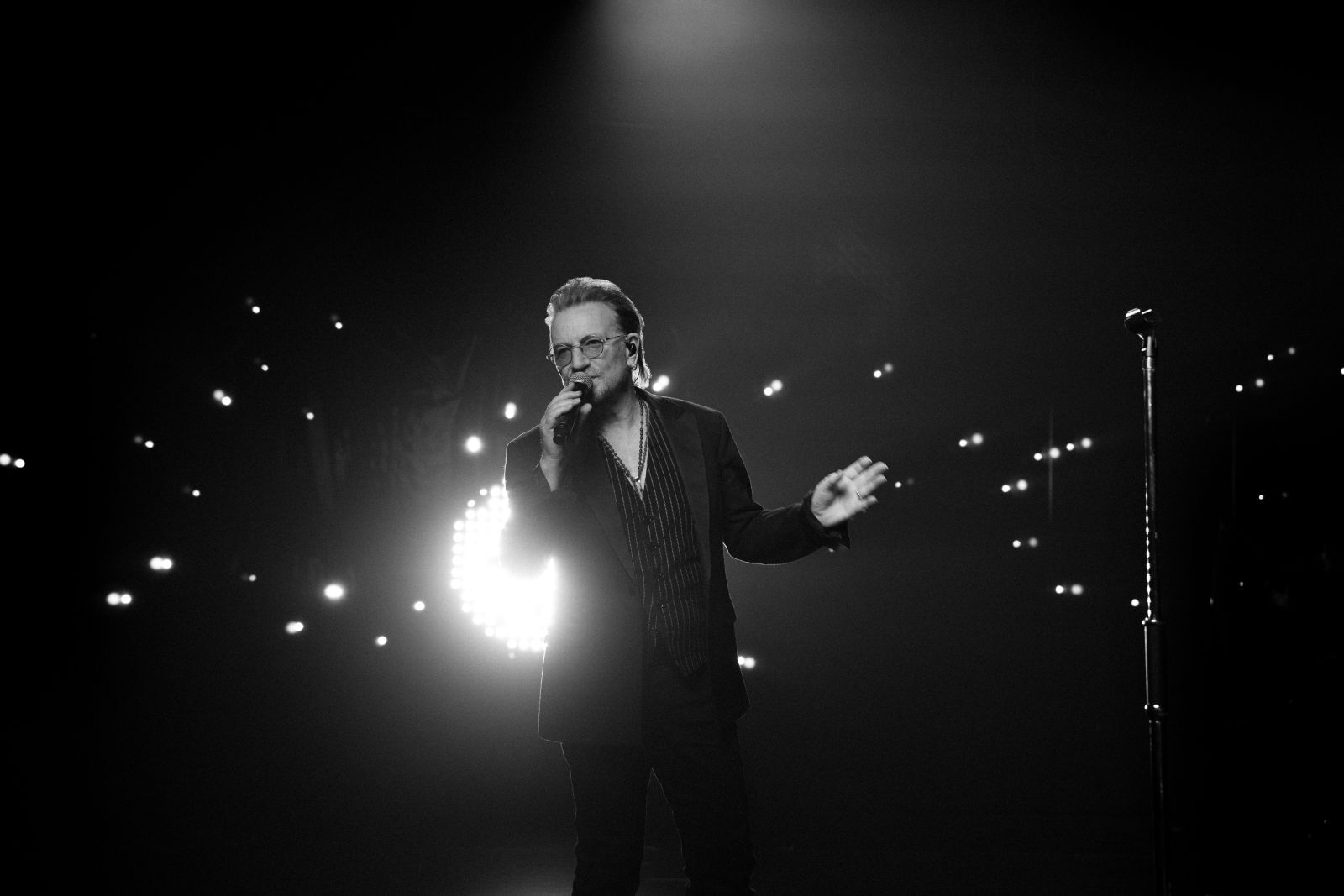 A black and white image of singer Bono standing on stage. He is wearing a black suite with sunglasses, and holding a microphone. There is a series of lights behind him.