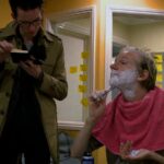 Julian Assange, a middle-aged white man with light blond hair, shaves his chin with a razor. A pink towel is draped over his shoulders and his face is covered with sharing cream. A person in a long olive green coat stands beside him and reads a book.