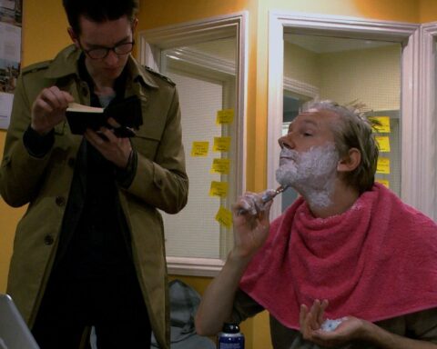 Julian Assange, a middle-aged white man with light blond hair, shaves his chin with a razor. A pink towel is draped over his shoulders and his face is covered with sharing cream. A person in a long olive green coat stands beside him and reads a book.