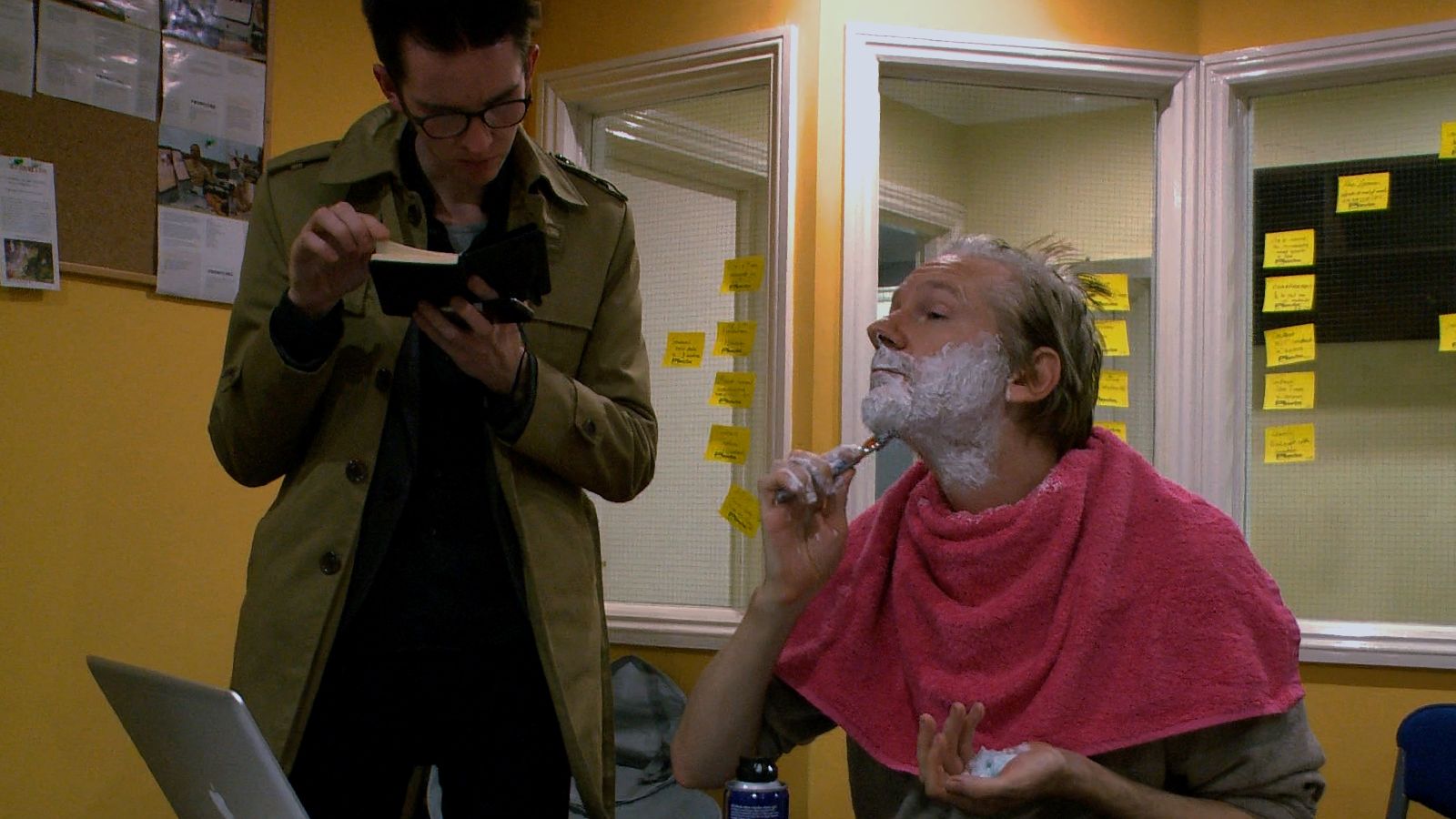 Julian Assange, a middle-aged white man with light blond hair, shaves his chin with a razor. A pink towel is draped over his shoulders and his face is covered with sharing cream. A person in a long olive green coat stands beside him and reads a book.