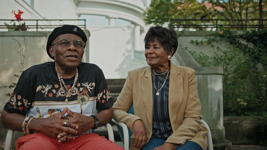 A Black couple--one man and one woman--sits together outdoors. The man is wearing a black and white t-shirt, red pants, sunglasses, a gold chain, and a black hat. The woman is wearing a tan blazer, striped shirt, and blue jeans. 