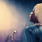 An animated image of Makayla Walker performing as Jackie Shane, a young Black transwoman. She is wearing a blue dress and a blonde wig. She is facing a spotlight.