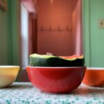 A watermelon sits in a red Pyrex bowl on a blue patterned table cloth. The melon is cut in half with its juicy pink flesh visible.