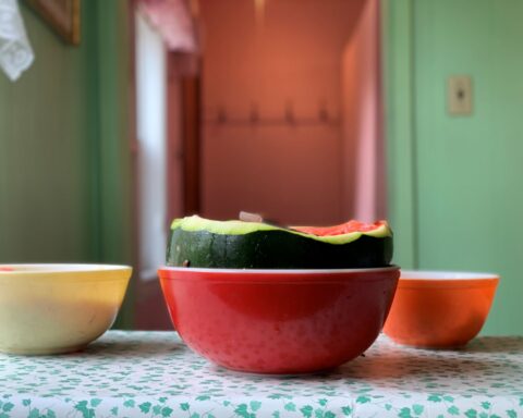 A watermelon sits in a red Pyrex bowl on a blue patterned table cloth. The melon is cut in half with its juicy pink flesh visible.