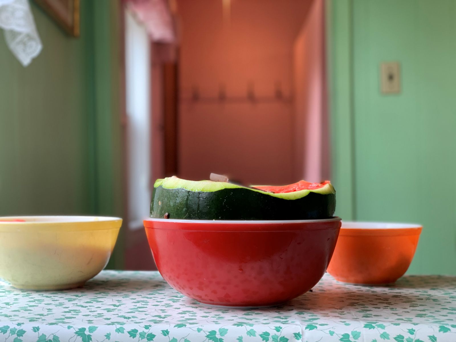 A watermelon sits in a red Pyrex bowl on a blue patterned table cloth. The melon is cut in half with its juicy pink flesh visible.