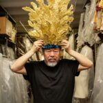 Chinese artist Ai Weiwei poses with a gold feathered headpiece atop his head. He is standing in a costuming department closet and wearing a black t-shirt. He has a bushy grey beard.