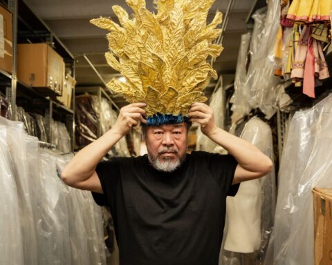 Chinese artist Ai Weiwei poses with a gold feathered headpiece atop his head. He is standing in a costuming department closet and wearing a black t-shirt. He has a bushy grey beard.