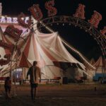 A boy approaches a circus tent at night. The words Circus Arena appear in red on an arch above the entrance.