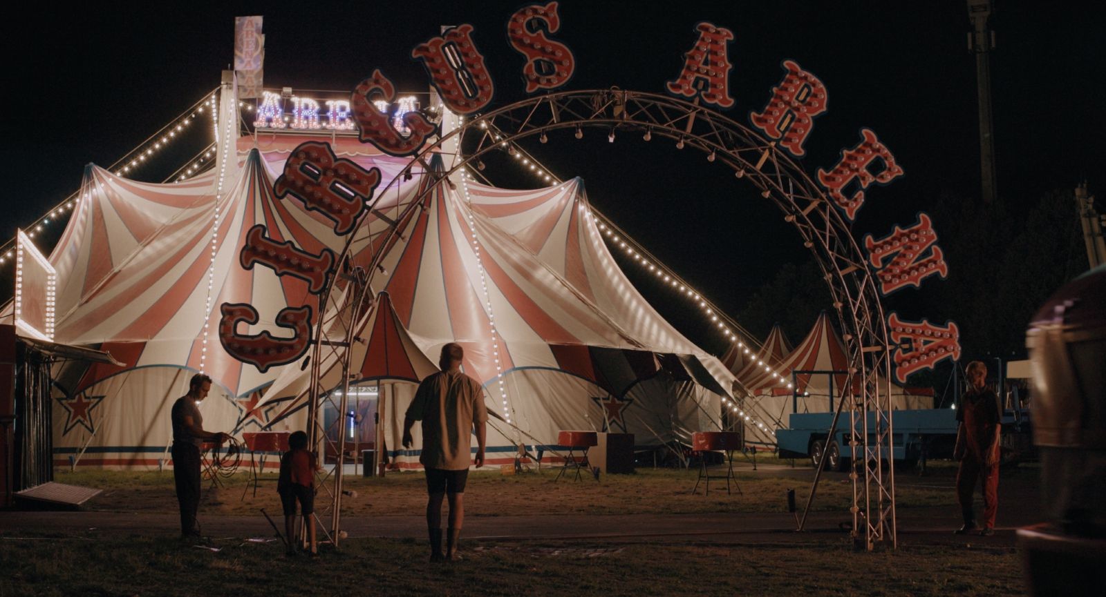 A boy approaches a circus tent at night. The words Circus Arena appear in red on an arch above the entrance.