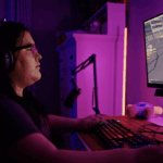 A young woman sits at a computer playing the game Roblox. She is wearing a black t-shrt, has dark brown hair, and is wearing glasses. Her room is lit by pink lights.