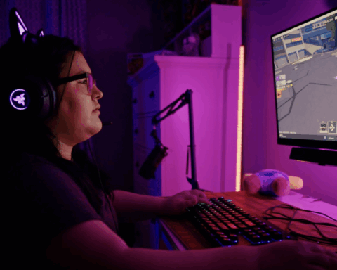A young woman sits at a computer playing the game Roblox. She is wearing a black t-shrt, has dark brown hair, and is wearing glasses. Her room is lit by pink lights.