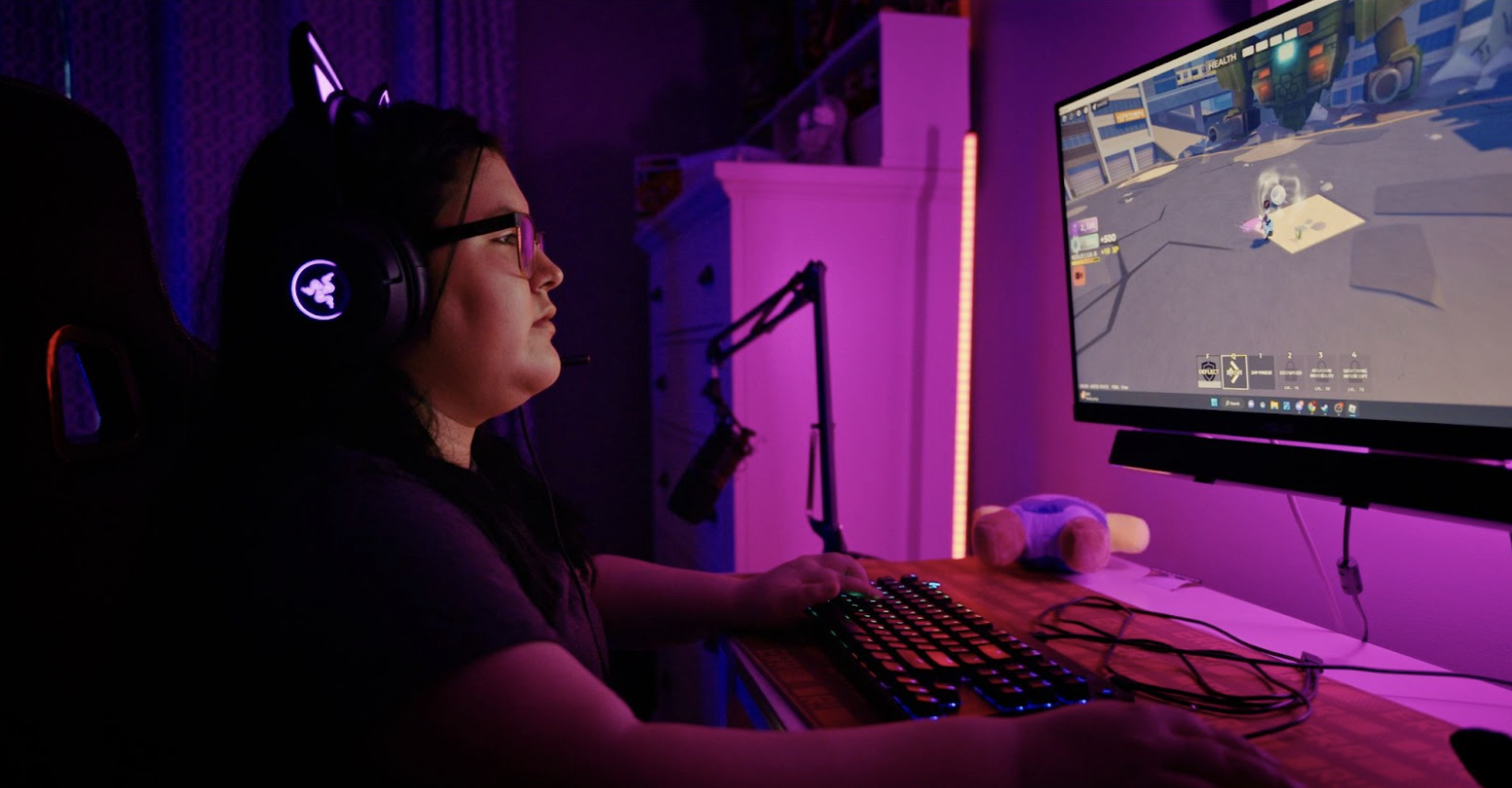 A young woman sits at a computer playing the game Roblox. She is wearing a black t-shrt, has dark brown hair, and is wearing glasses. Her room is lit by pink lights.