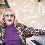 Amanda Lear sits at a piano. She is wearing a purple shirt with a leopard print blazer and blue-tinted glasses. She is a white woman with blond hair. One hand is raised to her face and she is laughing.