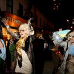 A drag queen leads a parade in the streets during Purim. She is wearing a black jacket and white dress, with a blonde wig, sunglasses, and festive attire.