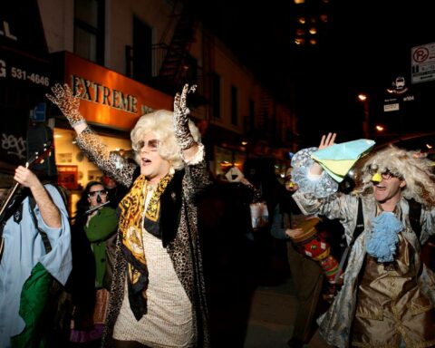 A drag queen leads a parade in the streets during Purim. She is wearing a black jacket and white dress, with a blonde wig, sunglasses, and festive attire.