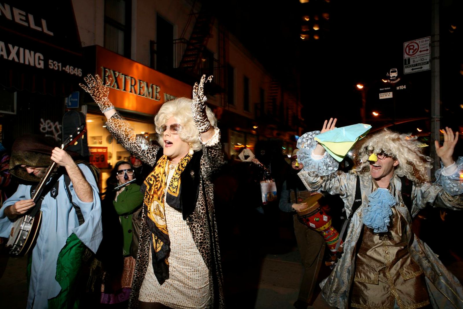 A drag queen leads a parade in the streets during Purim. She is wearing a black jacket and white dress, with a blonde wig, sunglasses, and festive attire.