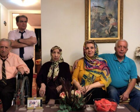 An aging Iranian family poses for a portrait. Two elderly parents sit on screen left with the father in a wheelchair and the mother seated beside her daughter and son-in-law. The son stands behind the father with his arms crossed.