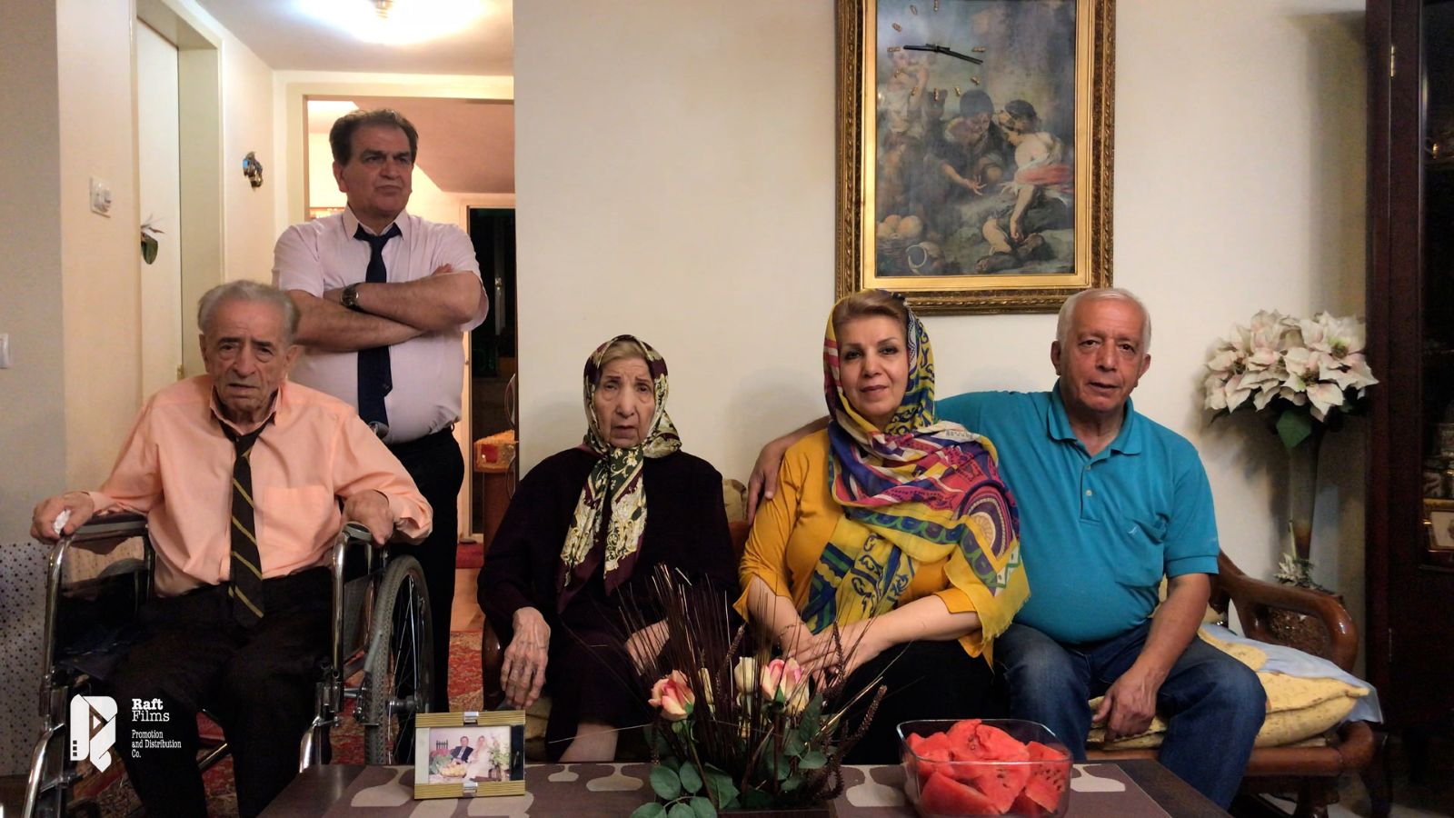 An aging Iranian family poses for a portrait. Two elderly parents sit on screen left with the father in a wheelchair and the mother seated beside her daughter and son-in-law. The son stands behind the father with his arms crossed.