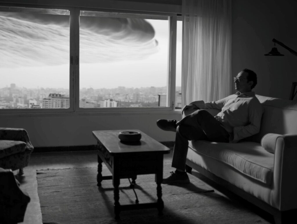 A black and white photo of a man sitting in his apartment. There is a large ominous cloud hanging over the city, as seen through the window behind him.