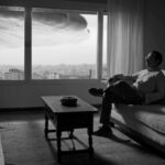A black and white photo of a man sitting in his apartment. There is a large ominous cloud hanging over the city, as seen through the window behind him.