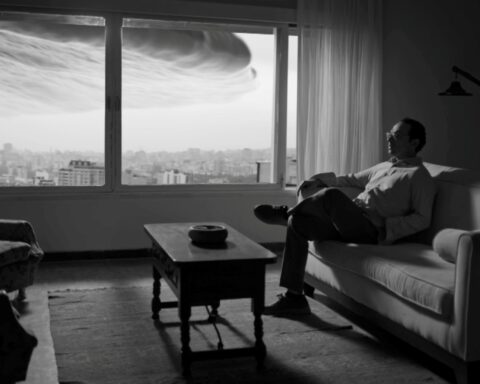 A black and white photo of a man sitting in his apartment. There is a large ominous cloud hanging over the city, as seen through the window behind him.