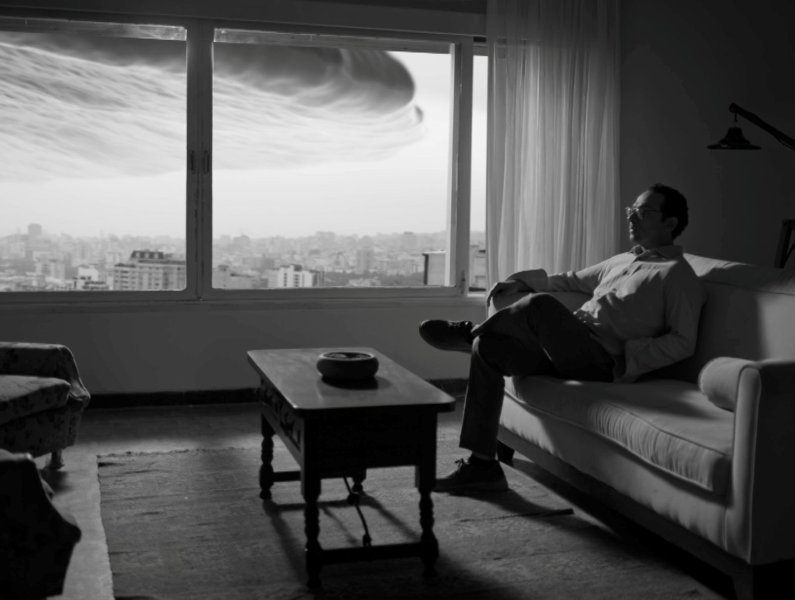 A black and white photo of a man sitting in his apartment. There is a large ominous cloud hanging over the city, as seen through the window behind him.