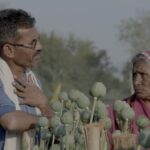 An Indian man and his elderly mother stand in a poppy field. The flowers have not yet bloomed. He has his hand on his chest while speaking to her.