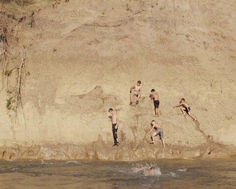 Five young boys play on a rocky shoulder near a pond.