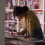 A young South Korean woman stands above a desk looking through stacks of photo albums. She is wearing a yellow sweater and there is a full bookcase behind her.