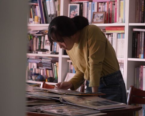 A young South Korean woman stands above a desk looking through stacks of photo albums. She is wearing a yellow sweater and there is a full bookcase behind her.