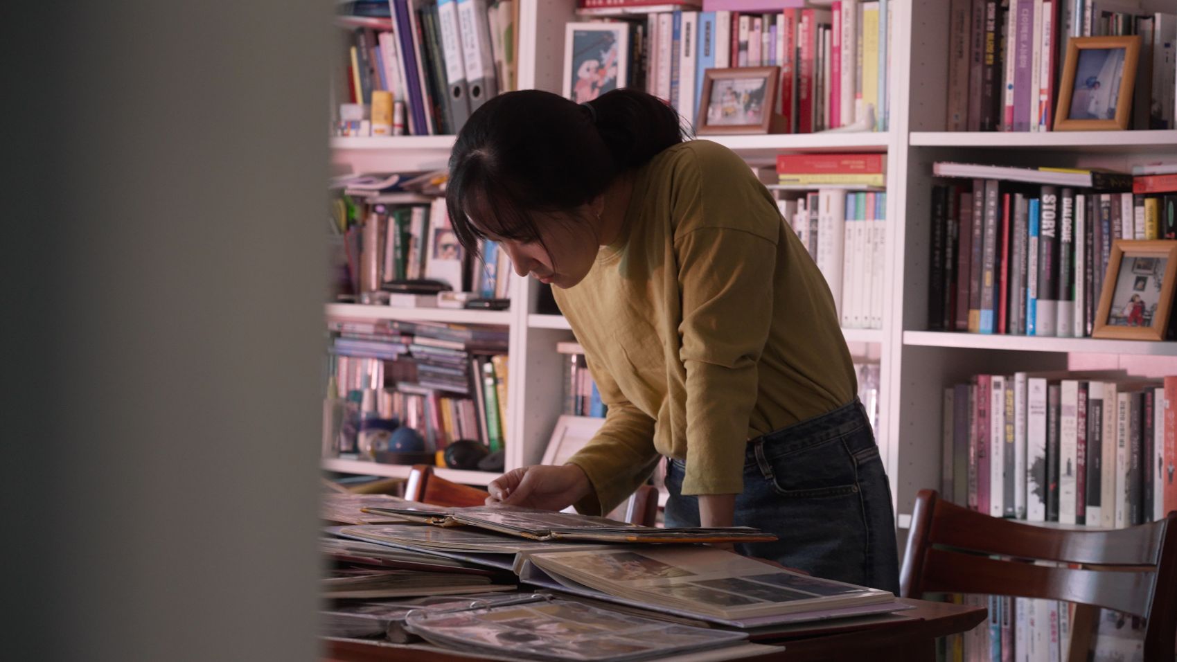 A young South Korean woman stands above a desk looking through stacks of photo albums. She is wearing a yellow sweater and there is a full bookcase behind her.