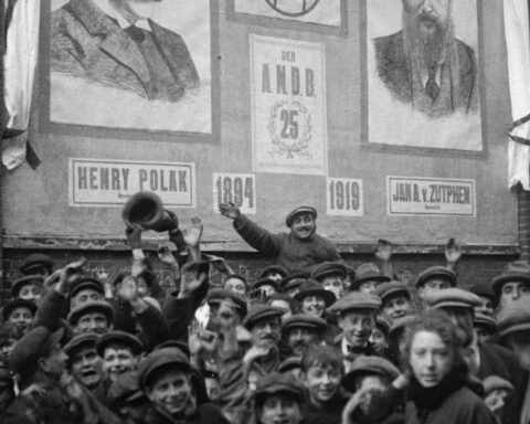 A black and white archival shot of a crowd in Amsterdam prior to World War II. A man in the middle is waving.