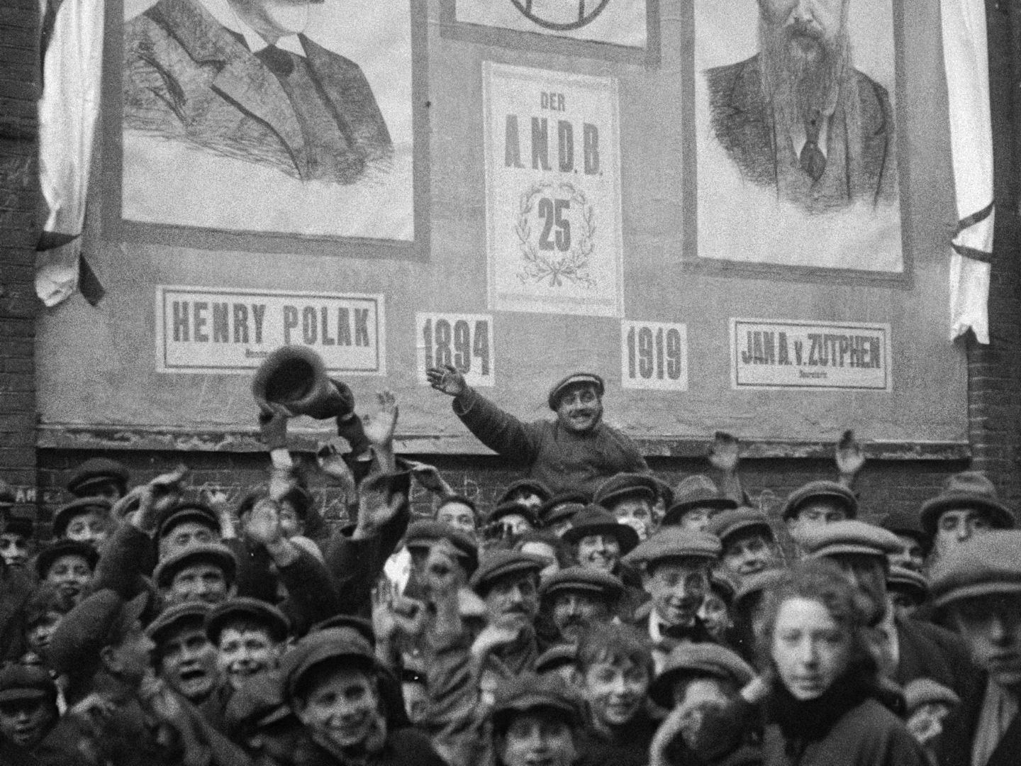 A black and white archival shot of a crowd in Amsterdam prior to World War II. A man in the middle is waving.